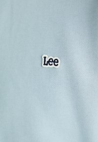 Tejido de algodón azul claro con una superficie texturizada, que presenta un pequeño logo bordado de "Lee" en azul marino y blanco en la parte superior izquierda.