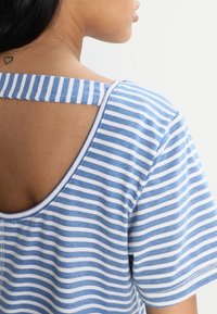 Chemise à rayures à manches courtes en coton bleu et blanc, avec un dos arrondi et un détail de sangle horizontale.