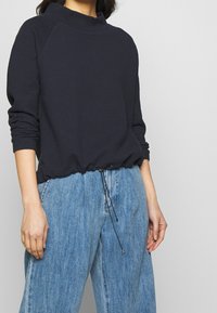 Marinblå texturerad tröja med hög krage och dragsko i nederkant, kombinerad med ljusblå vida jeansbyxor. Avslappnad passform, ledig stil.