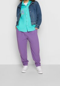 Denimjacka, turkos knappad skjorta med grafisk detalj och lösa lila byxor. Vita sneakers fulländar outfiten.