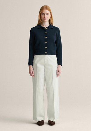 Rødhåret kvinde iført en sort cardigan med knapper, højtaljede, vide hvide bukser og brune sko, stående mod en ensfarvet beige baggrund.