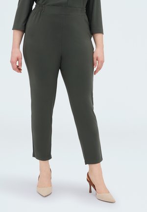 Kobieta nosi ciemnozielone, zwężane spodnie z elastycznym pasem i beżowe sandały typu slingback na obcasie z ostrym noskiem na jednolitym tle.