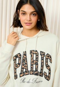 Grietas pūkinis su užrašu "PARIS" leopardų rašto raidėmis. Su kuoduku, elastinėmis rankovėmis ir minkšta tekstūra. Apačioje yra "Île-de-France" dizainas.