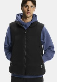 Chaleco acolchado negro con cuello alto, cremallera frontal y bolsillos laterales. Lleva puesto un sudadera azul con capucha. Presenta un logo en la parte inferior derecha.