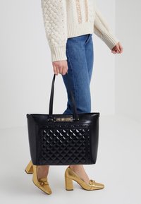 Black leather tote bag with quilted pattern, gold logo plaque, and dual handles. Model wears a knitted sweater and denim, holding the bag.