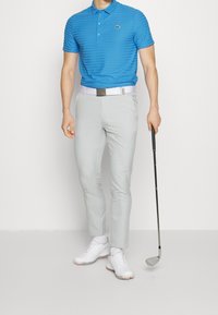 Blårandig polo skjorta, ljusgrå slim fit byxor, vita golfsandaler, och håller en golfklubba. Har ett bälte med spänne i metall.