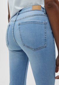 Jean en denim bleu clair à coupe ajustée, présentant deux poches arrière, des surpiqûres contrastées et un patch en cuir marron clair à la ceinture.