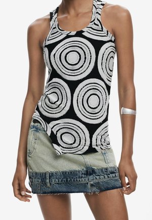 Femme portant un haut noir sans manches avec de grands motifs circulaires blancs et une jupe en denim bicolore courte, bracelet argenté sur le bras gauche.