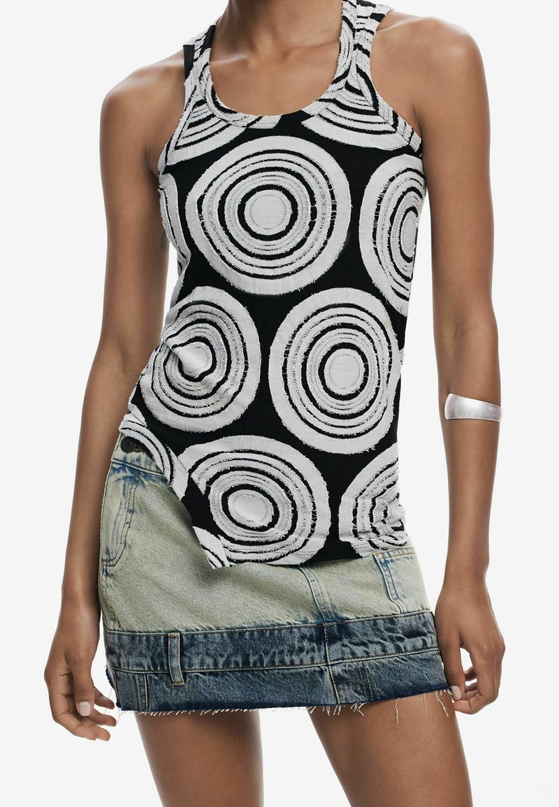 Femme portant un haut noir sans manches avec de grands motifs circulaires blancs et une jupe en denim bicolore courte, bracelet argenté sur le bras gauche.