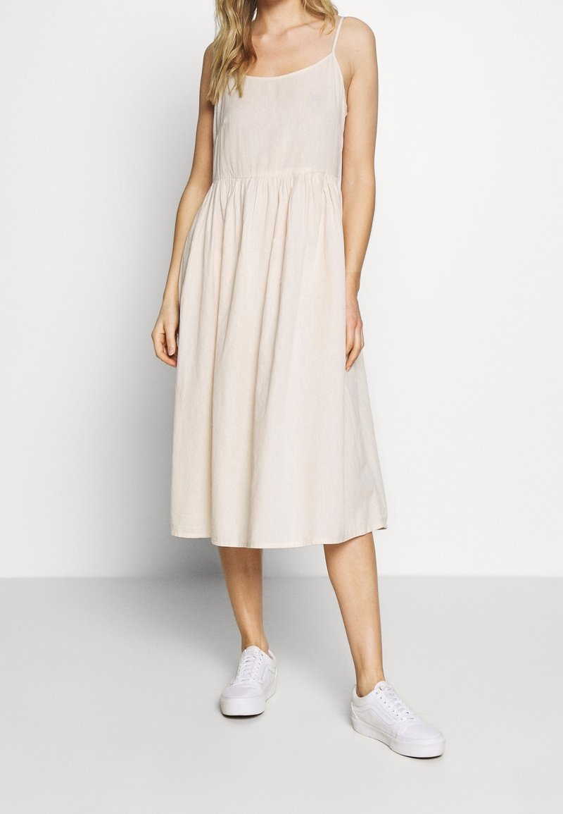 Beżowa bawełniana sukienka na ramiączkach z cienkimi strapsami, zebranym w pasie designem i zwiewnym dołem, w połączeniu z białymi trampkami. Gładka faktura i luźny krój.