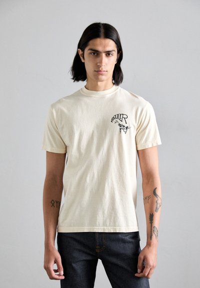 Tricou crem din bumbac, cu mâneci scurte, guler rotund și imprimeu grafic negru pe partea stângă a pieptului; purtat cu blugi din denim închis.