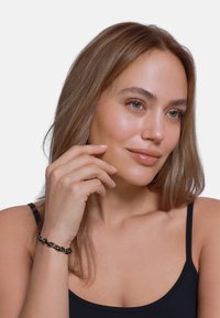 Schwarzes geflochtenes Armband auf einem Handgelenk präsentiert, mit einem Kettenmuster und goldenen Akzenten. Glatte Hautfarbe, hellbraunes Haar, neutrales Oberteil.
