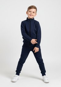 Granatowy rozpinany sweter z wysokim kołnierzem i pasujące spodnie. Sweter ma ściągacze na mankietach, w połączeniu z białymi trampkami. Gładka faktura.