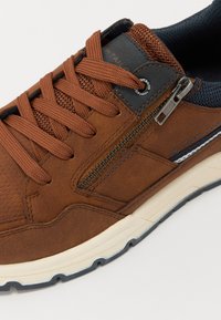 Sneaker en cuir brun avec des accents en textile, présentant un système de laçage, une fermeture éclair latérale et des détails texturés sur le dessus. Semelle extérieure en caoutchouc.
