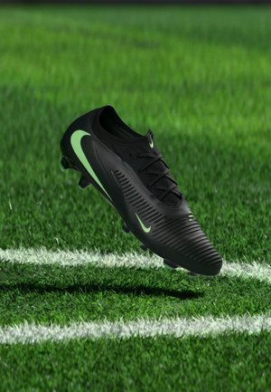 Schwarzer Fußballschuh mit grünem Nike-Swoosh schwebt über einem Grasfeld nahe weißen Linienmarkierungen.