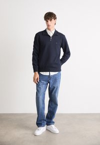 Navy zip-up sweatshirt with ribbed cuffs and neck, layered over a white collared shirt, paired with light blue denim and white sneakers.