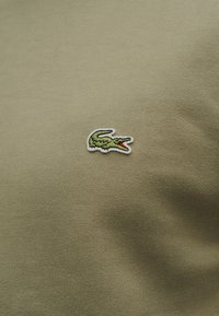 Logo de crocodile vert brodé sur un tissu vert olive, centré sur la poitrine d'un vêtement.