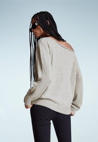 Frau mit langen Zöpfen, die schwarze Sonnenbrillen trägt, einen hellgrauen schulterfreien Pullover und schwarze Hosen anhat, steht vor einem schlichten hellen Hintergrund.