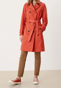 Červený trenčkot s páskem, dvouřadovým zapínáním a černými knoflíky. Kombinován s béžovými kalhotami a červenými teniskami s bílou podrážkou.