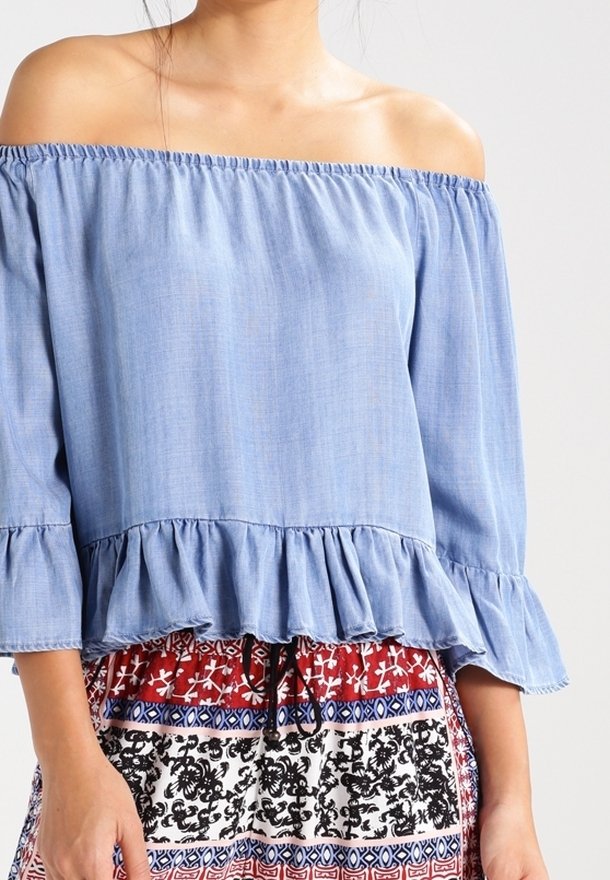 Blusa azzurra chiara con scollatura a spalle scoperte, colletto elasticizzato e orlo arricciato. Maniche lunghe con polsini arricciati. Tessuto morbido al tatto.