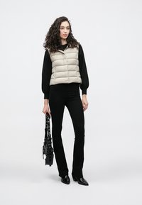 Beige puffer vest over a black sweater, paired with black flared pants. The model holds a black bag with textured details.