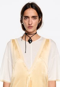 Abito in raso giallo pallido con scollatura a V profonda e bordo in pizzo, sovrapposto a una camicia bianca; presenta un ciondolo a forma di cuore nero su una collana di corda.