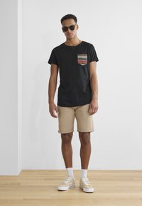 Svart t-skjorte med kort erme og mønstrete lomme, khaki shorts og beige sneakers. Modellen har solbriller; bakgrunnen er minimalistisk.