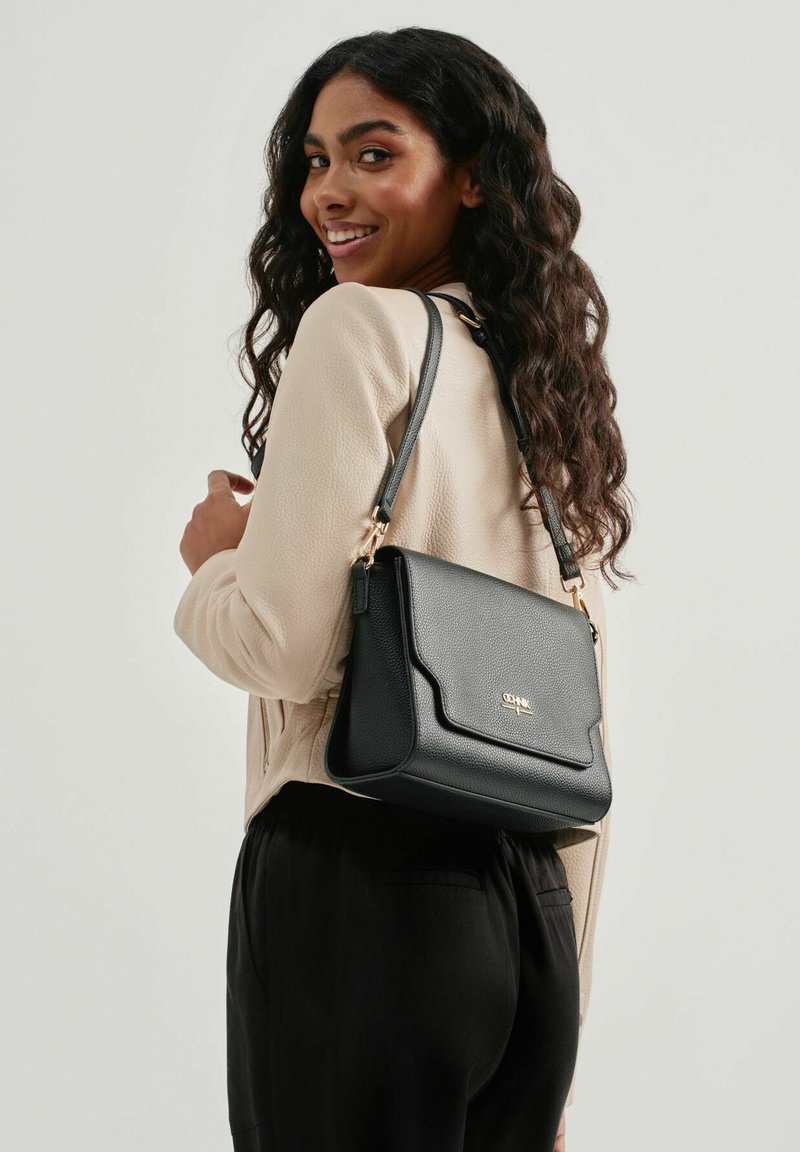 Czarna torebka ze skóry z fakturą w drobne kamyki, z klapką, złotymi akcentami i regulowanym paskiem, noszona na ramieniu na tle jasnej powierzchni.