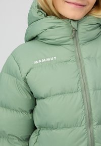 Lichtgroene pufferjack met hoge kraag, frontale rits en opvallende stiksels. Kenmerkt zich door het witte "Mammut" logo op de borst.