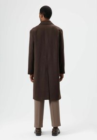 Bruine lange jas met een rechte snit, een inkeping in het revers en een rugsplit. Gedragen over lichtbruine broek en donkere schoenen, tegen een neutrale achtergrond.