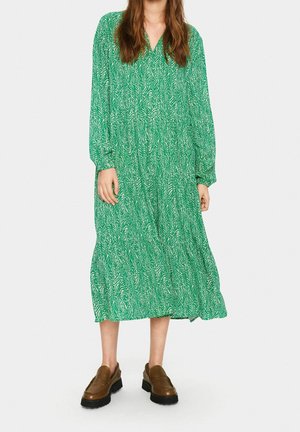 Robe midi verte avec un motif de feuilles, à manches longues et décolleté en V. Associée à des chaussures brunes à enfiler et à un design superposé et fluide.