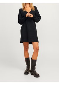 Abito nero a maglia con scollatura a V, maniche lunghe e vestibilità ampia. Abbinato a stivali lucidi neri al ginocchio. Sfondo semplice.