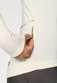 Langsleeve, crèmekleurig top met een textuurstof, voorzien van een zijzak met een roze ritssluiting. Naadtoeslag is zichtbaar nabij de zoom.