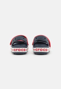 Crvene i mornarsko plave Crocsice s oblikovanim dizajnom, otvorenom stražnjom stranom i bijelim detaljima. Imaju "crocs" logo na peti. Teksturirana unutrašnjost.