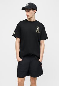 Νέος άνδρας που φοράει μαύρο καπέλο, μαύρο μπλουζάκι με μικρό γραφικό αστροναύτη και μαύρο σορτς με τσέπες, στέκεται με τα χέρια στις τσέπες.