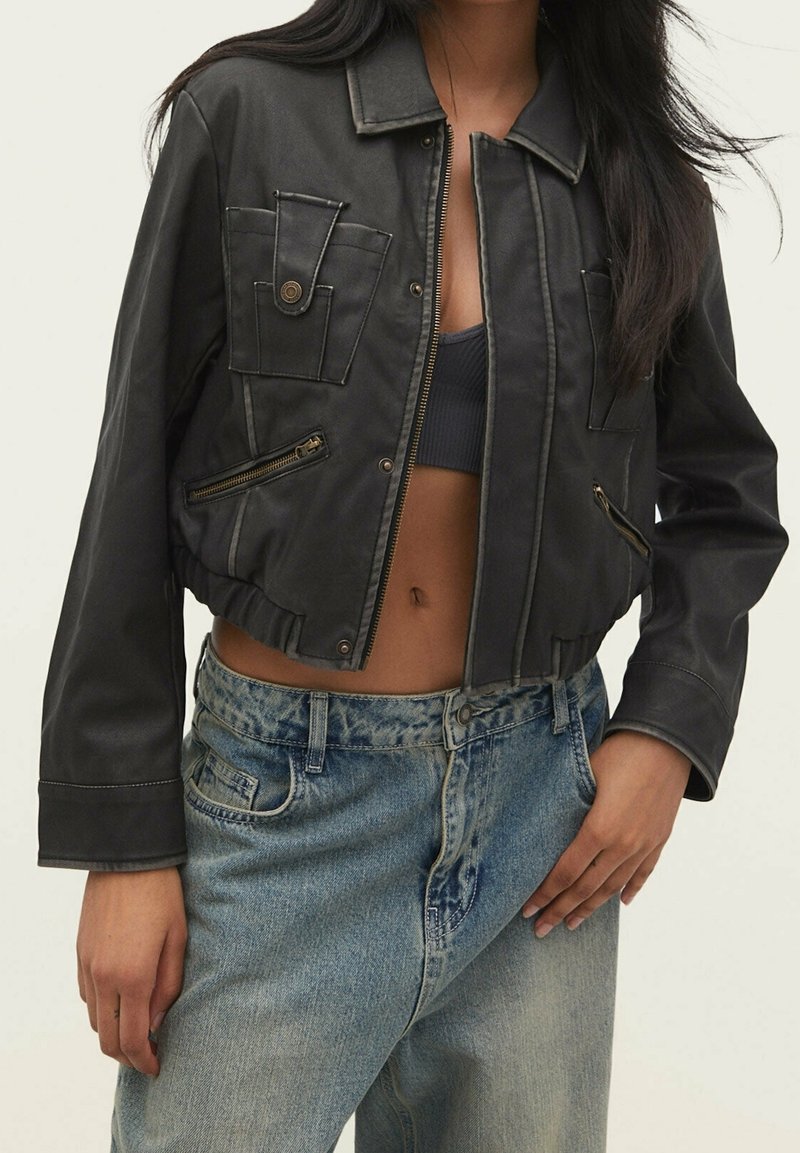 Zwarte cropped jas met voorzakken, ritssluitingen en een kraag. Gecombineerd met lichtblauwe spijkerbroek met een ontspannen pasvorm.