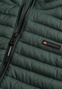 Grön quiltad jacka med en svart dragkedja, med en texturerad tyg och en liten svart Superdry-logotyp på bröstet.
