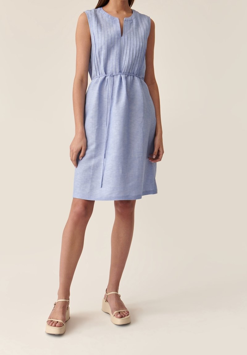 Robe bleu clair sans manches avec corsage plissé, encolure en V et taille à cordon. Le modèle porte des sandales compensées beiges.