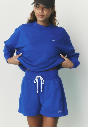 Nuori nainen, jolla on sininen collegepaita ja siihen sopivat shortsit, joissa on teksti "self.", säädellä collegepaitaa ja musta baseball-lippalakki yksiväristä taustaa vasten.