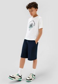 Weißes T-Shirt mit Palmen-Graphic, marineblaue Shorts und schwarz/weiße Sneakers mit grünen Akzenten. Die Textur wirkt weich; das Design ist lässig und sportlich.