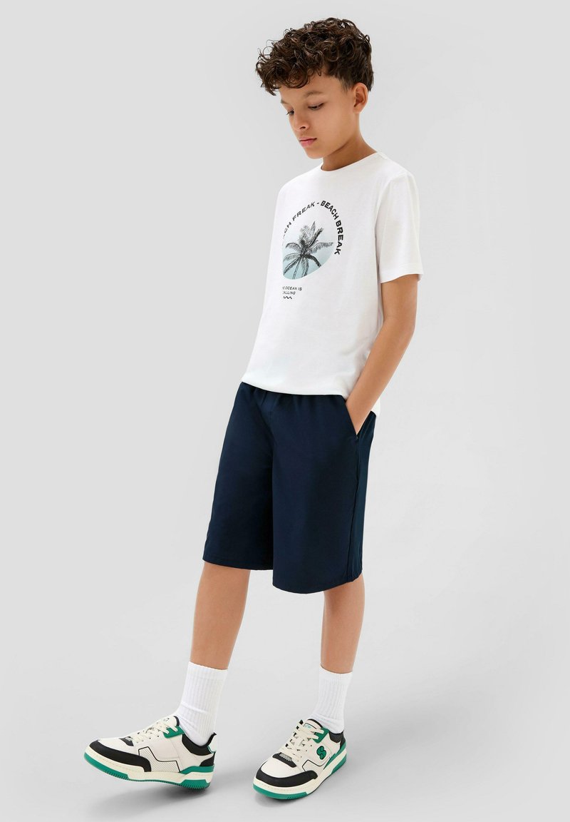 Weißes T-Shirt mit Palmen-Graphic, marineblaue Shorts und schwarz/weiße Sneakers mit grünen Akzenten. Die Textur wirkt weich; das Design ist lässig und sportlich.
