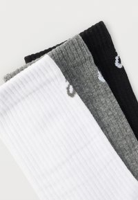 Τρία ζεύγη διπλωμένων ριγωτών κάλτσες σε λευκό, γκρι και μαύρο, με ένα μικρό λογότυπο κοντά στις μανσέτες, τοποθετημένα επικαλυπτόμενα σε μια λευκή επιφάνεια.