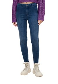 Žena v tmavě modrých skinny džínách, fialovém pleteném svetru a bílých teniskách do vysokého střihu stojí před bílým pozadím.