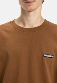 Brązowa bawełniana koszulka z okrągłym dekoltem i krótkimi rękawami, z czarno-białym haftowanym logo po lewej stronie klatki piersiowej.