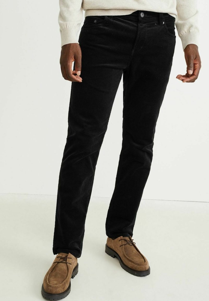 Personne portant un pantalon noir ajusté et des chaussures à lacets en daim marron, debout sur un sol clair.