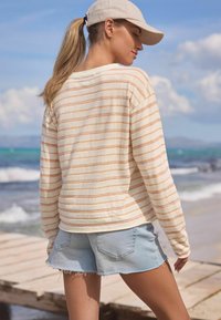 Stripet langærmet top i creme med orange striber, parret med lyseblå denimshorts med en flosset kant, sat op mod en strandbaggrund.