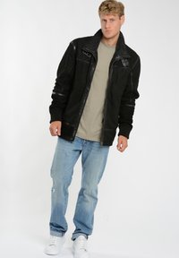 Sort ruskind jakke med shearling krave, lynlås lukning, tekstureret krop og polstrede skuldre, båret over en lys grøn skjorte og blå jeans.