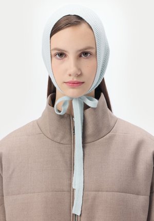 Jeune femme portant un bonnet en tricot bleu clair noué sous le menton, vêtue d'une veste beige zippée, sur un fond uni.