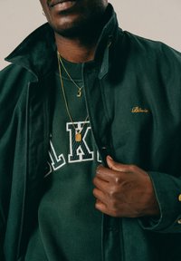 Άνδρας με σκούρο πράσινο μπουφάν με χρυσές κεντητές λεπτομέρειες, πράσινη μπλούζα με λευκά γράμματα και δύο χρυσά κολιέ, που κρατάει τον γιακά του μπουφάν.