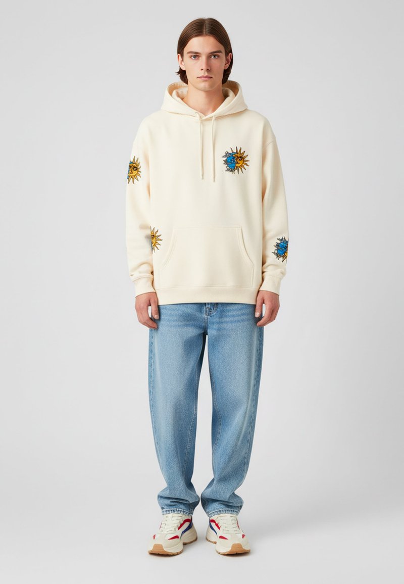 Sudadera crema con bordado de sol y luna, bolsillo canguro, combinada con pantalones vaqueros azul claro y zapatillas de colores. Tejido suave, corte relajado.